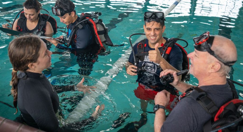 Individuals wearing swimsuits and scuba gear in a pool. One man smiles and offers a thumbs up.